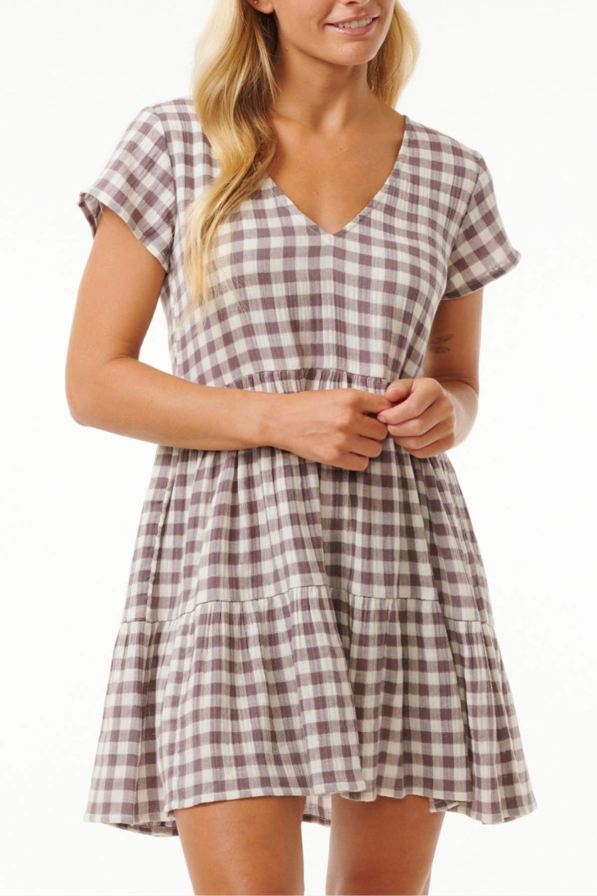 A woman is dressed in a short-sleeved, V-neck Rip Curl Premium Surf Check Dress featuring a tiered skirt and a white and brown checkered pattern. The dress, made from cotton double weave gauze with yarn-dyed fabric, matches her relaxed smile as she gently rests her hands by her side.
