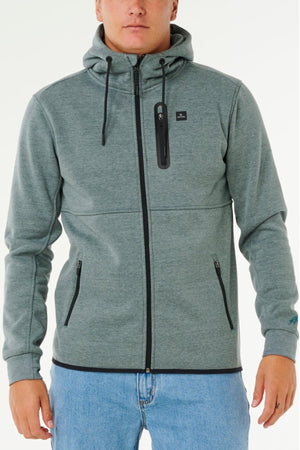 A person wears a light gray Rip Curl Anti Series Departed Zip Thru hoodie with black drawstrings and zipper, paired with light blue jeans, standing against a plain white background for a stylish winter look.