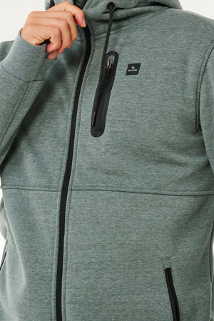 A person wears a gray Rip Curl Anti Series Departed Zip Thru hoodie with black zippers and a small chest logo, holding the hood near their face. Only their upper body is visible, highlighting this stylish winter garment.