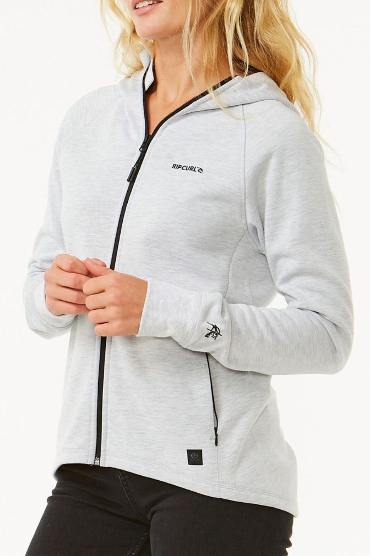 A woman wearing a light gray Rip Curl Anti Series Flux III Jacket with a hood, black zipper, and chest logo stands against a white background. This lightweight, shower-resistant jacket offers both style and function.