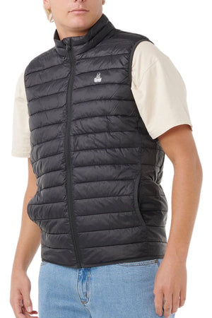 Someone stands against a plain white background, wearing the Rip Curl Anti Series Search Puffer Vest—black, water repellent, and lightweight—over a beige short-sleeve t-shirt and blue jeans to showcase practical style and warmth.