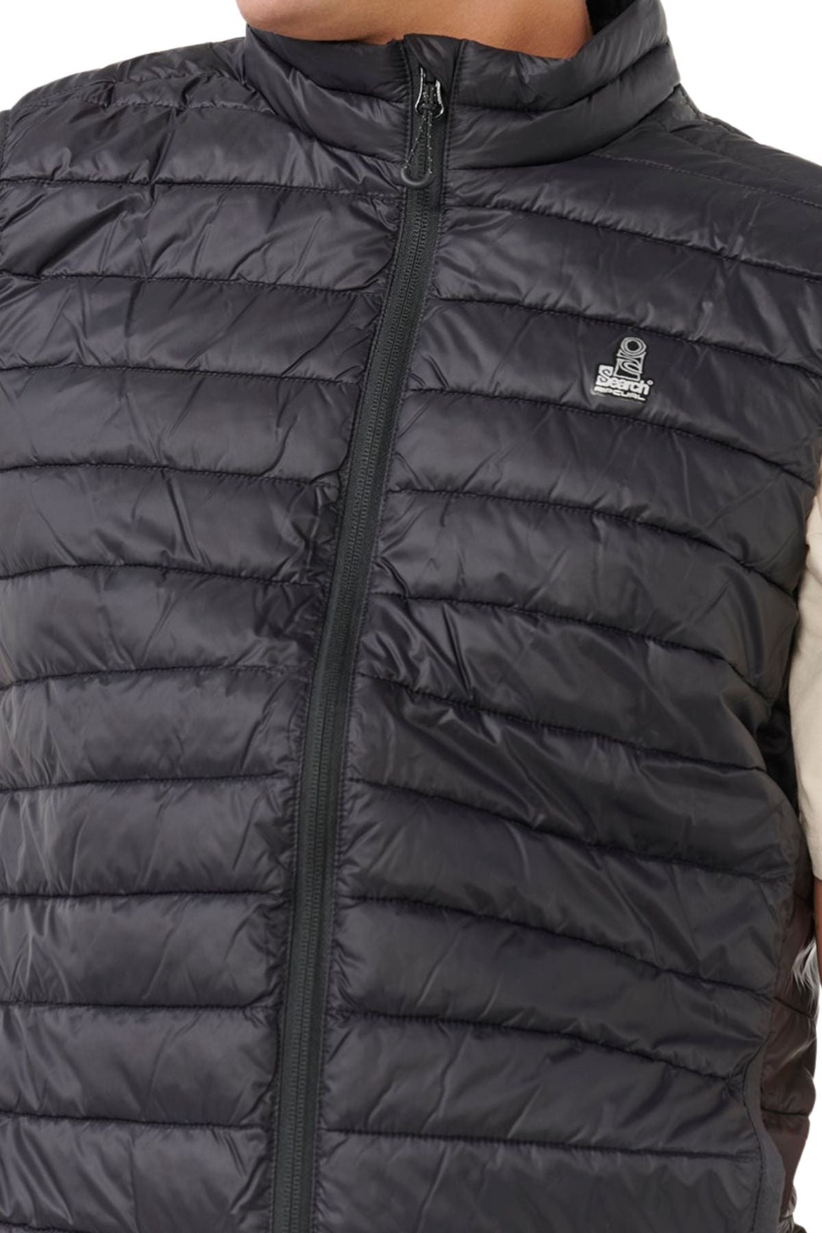 A close-up shows a person wearing the Rip Curl Anti Series Search Puffer Vest, offering lightweight warmth. The black vest features a small logo on the left chest and a center zipper, with only the torso and part of an arm visible.