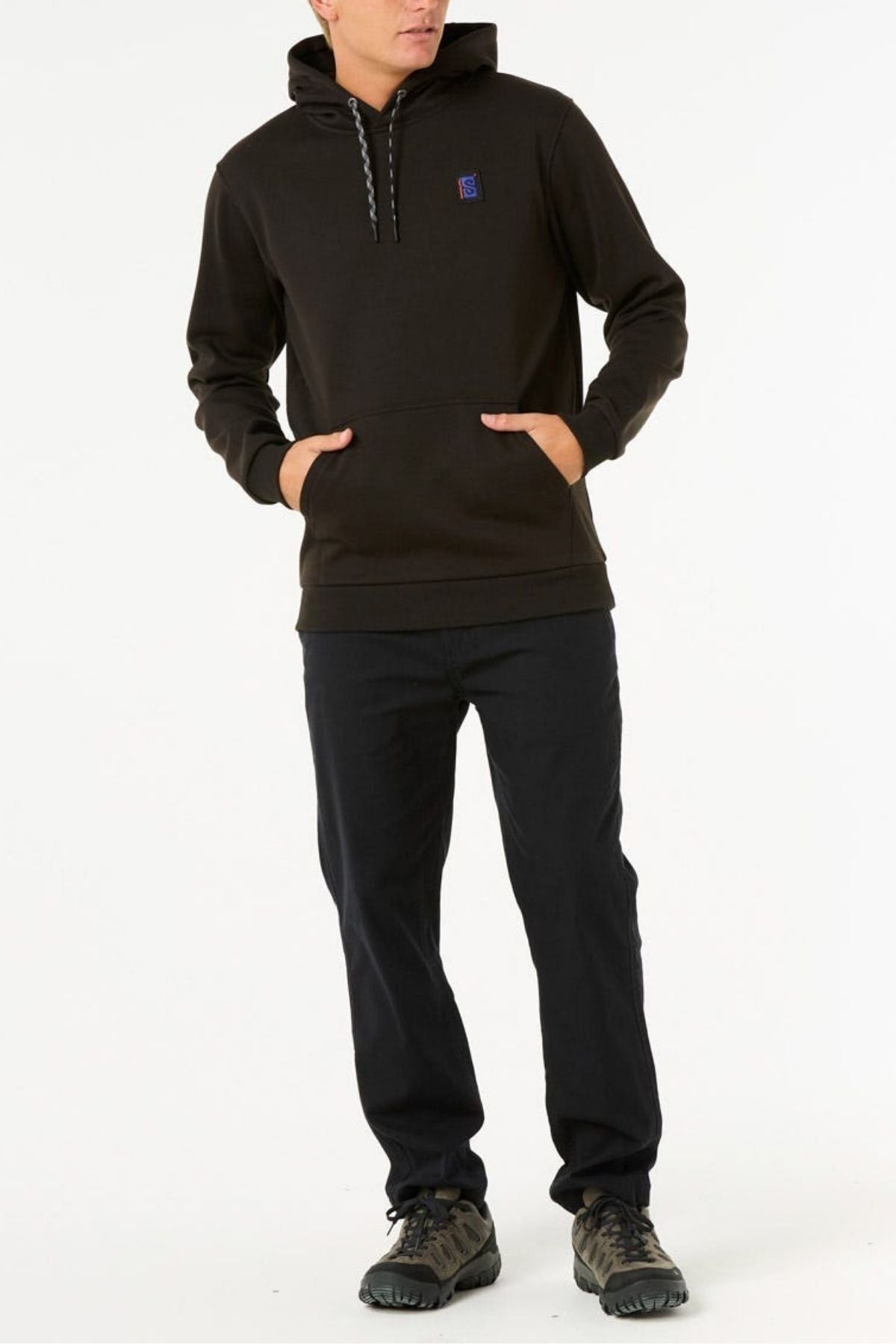 Someone wearing a Rip Curl Anti Series Search Base Hood in black with a front pocket and matching black pants stands with hands in the hoodie pocket against a plain white background.
