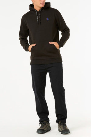 Someone wearing a Rip Curl Anti Series Search Base Hood in black with a front pocket and matching black pants stands with hands in the hoodie pocket against a plain white background.