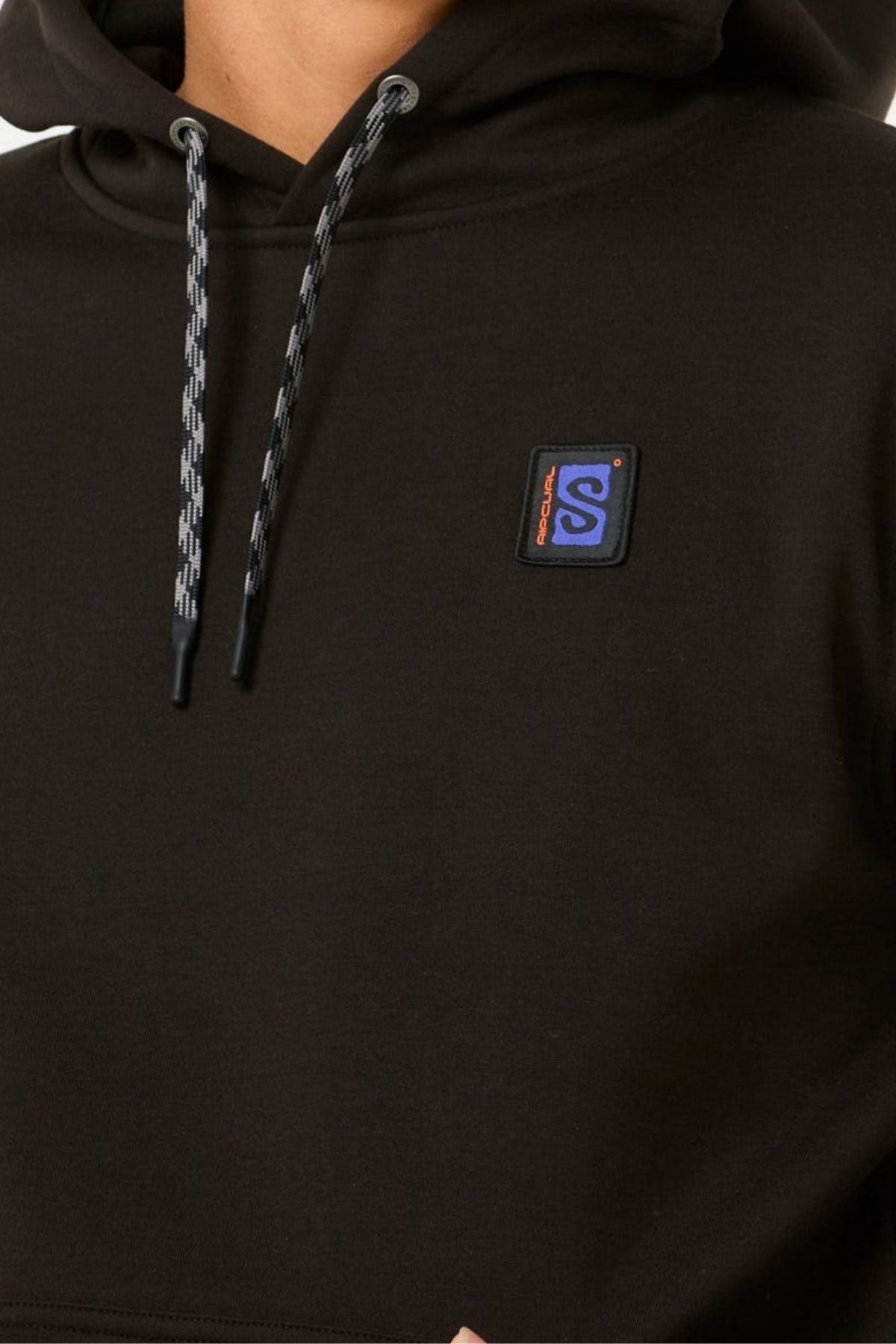 A close-up of a person wearing the Rip Curl Anti Series Search Base Hood in black, featuring patterned drawstrings, water repellent fabric, and a small blue and orange square logo patch on the chest.