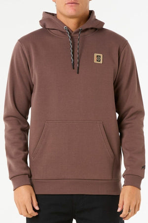 A person wears the Rip Curl Anti Series Search Base Hood—a brown hoodie with a bonded polar fleece lining, front pocket, and small square logo on the left chest—while standing against a plain white background.
