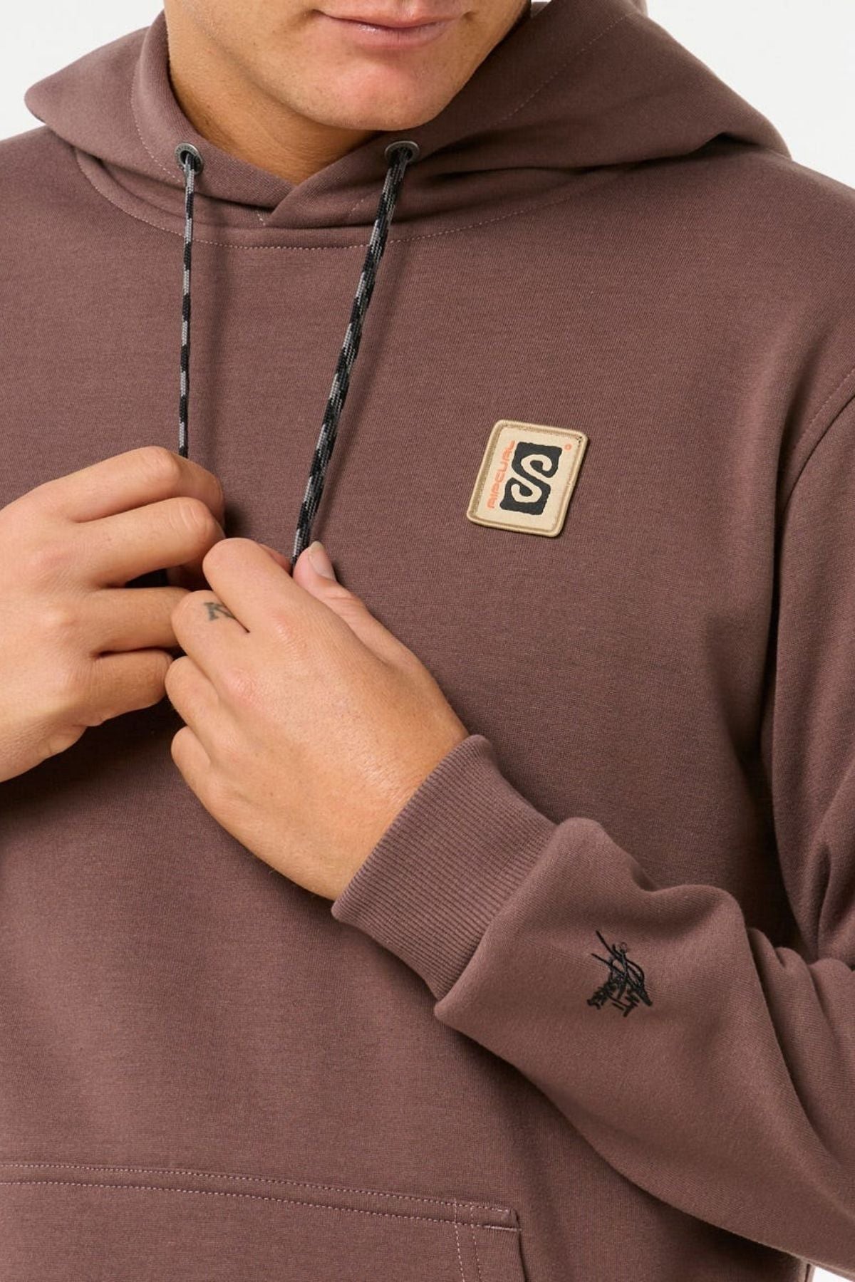 Wearing the Rip Curl Anti Series Search Base Hood—a brown, standard fit hoodie with an "S" chest patch, small black sleeve logo, and drawstrings—this person’s lower face is visible. Made from bonded polar fleece for extra warmth.