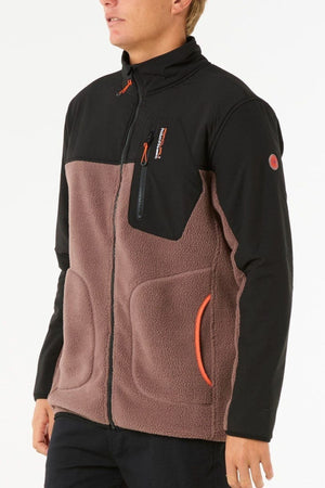 A person wearing the Rip Curl Anti Series Search Zip Crew—a two-tone polyester polar fleece jacket with a black top, brown lower section, orange zippers, and side pockets—stands against a plain white background.