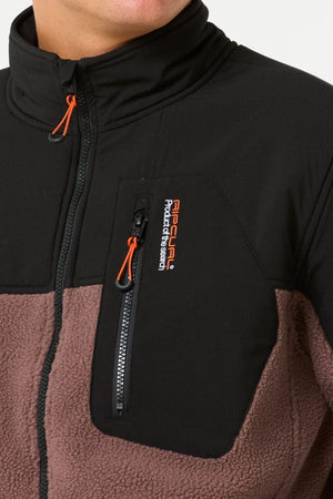 Close-up of a person wearing the Rip Curl Anti Series Search Zip Crew, a two-tone black and brown polyester fleece with a high collar. The chest pocket has an orange zip pull, Rip Curl logo, and “Product of the Search” text.