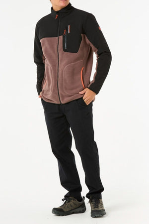 A person stands against a white background wearing a black and brown Rip Curl Anti Series Search Zip Crew polyester fleece jacket, dark pants, and gray hiking shoes with one hand in their pocket. Their face is not visible.