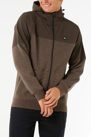 Wearing the Rip Curl Anti Series Viral Zip Thru—a brown zip-up hoodie with a hood and black zipper—a smiling person stands against a white background, hands loosely clasped; it’s the perfect stylish adventure wear.