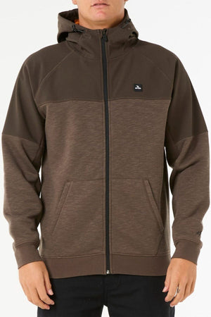 A person wearing a brown Rip Curl Anti Series Viral Zip Thru hoodie with a small chest logo, standing against a plain white background—ideal for anyone wanting stylish adventure wear.