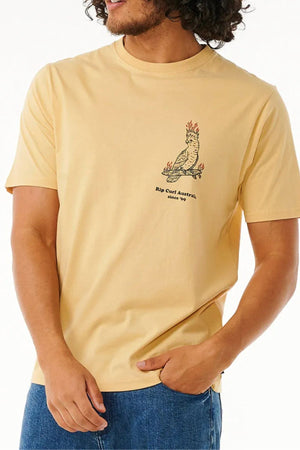 A man wears the Rip Curl Australia Fun Times Desto Tee, a light yellow cotton t-shirt featuring a small flaming bird graphic and "Rip Curl Australia, since 1969" text on the left chest, paired with blue jeans.