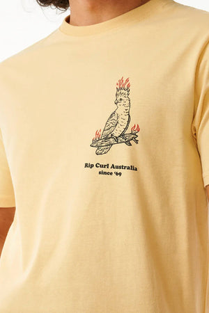 Someone wearing the Rip Curl Australia Fun Times Desto Tee—a light yellow cotton T-shirt featuring a flaming-head cockatoo on a branch and "Rip Curl Australia since '69" printed below the illustration.
