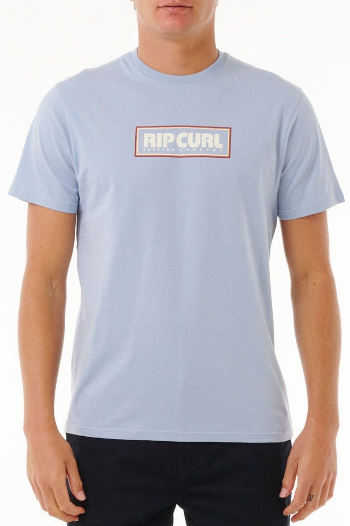 A person is wearing a casual Rip Curl Big Mumma Icon Tee in light blue with the logo prominently displayed. The organic cotton shirt is stylishly paired with black pants against a plain white background.