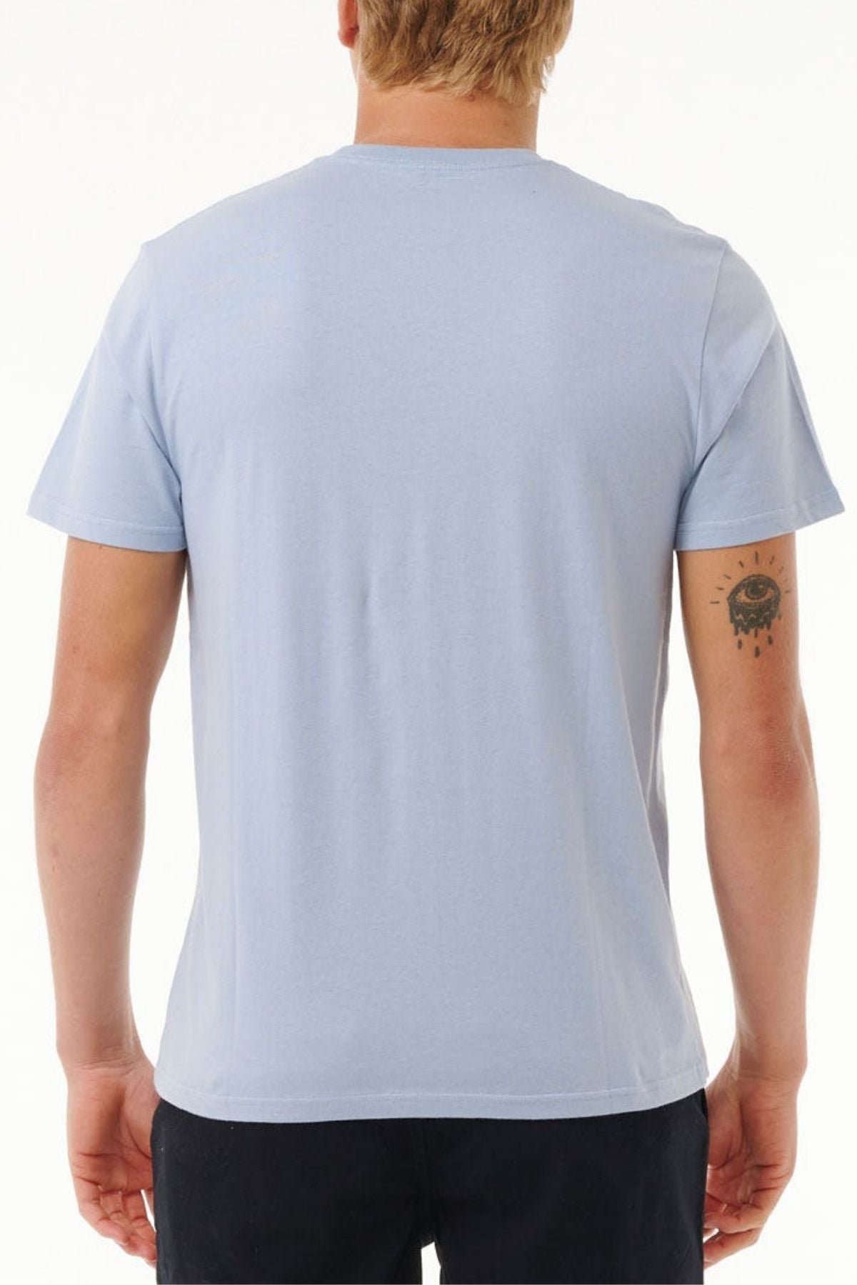 A person with short blonde hair is facing away, wearing a light blue Rip Curl Big Mumma Icon Tee and black pants. An eye tattoo with tears adorns their left arm, showcasing effortless style against a plain white background.