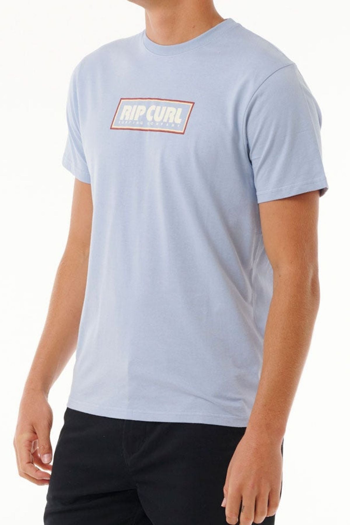 Someone is wearing a light blue Rip Curl Big Mumma Icon Tee made from organic cotton, featuring a "RIP CURL" logo in a red-bordered rectangle on the chest. They're against a plain white background, with face hidden, short sleeves worn, and paired with black trousers.