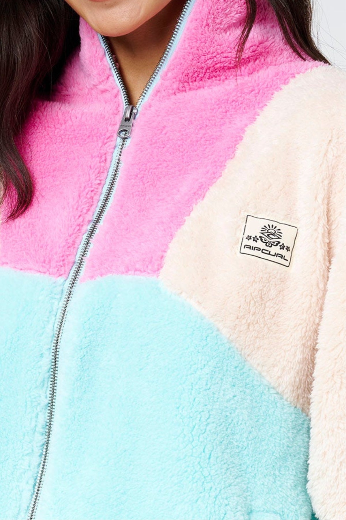 Wearing the Rip Curl Block Party II Zip Through Polar Fleece in pink, cream, and light blue with a chest sun patch and front zipper—ideal for outdoor adventures. Only the lower face and upper torso are visible.
