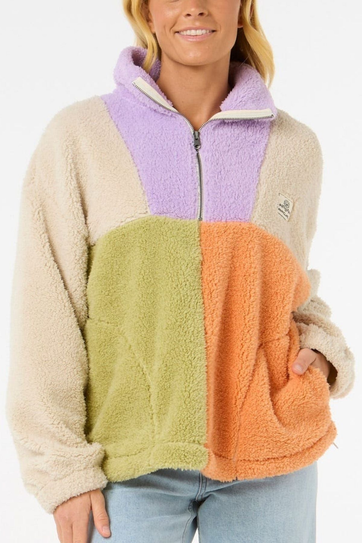 A woman wears the Rip Curl Block Party Polar Fleece, a cozy zip-up with cream, lavender, light green, and orange color blocks. It features a high collar, relaxed fit, and a unique look, paired here with light blue jeans.
