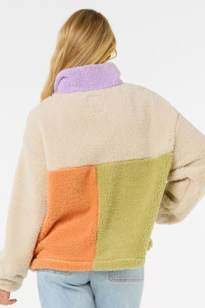 Wearing the Rip Curl Block Party Polar Fleece with its cozy, cream, orange, green, and purple color-blocked design and light blue jeans, a long-haired blonde individual faces away from the camera.