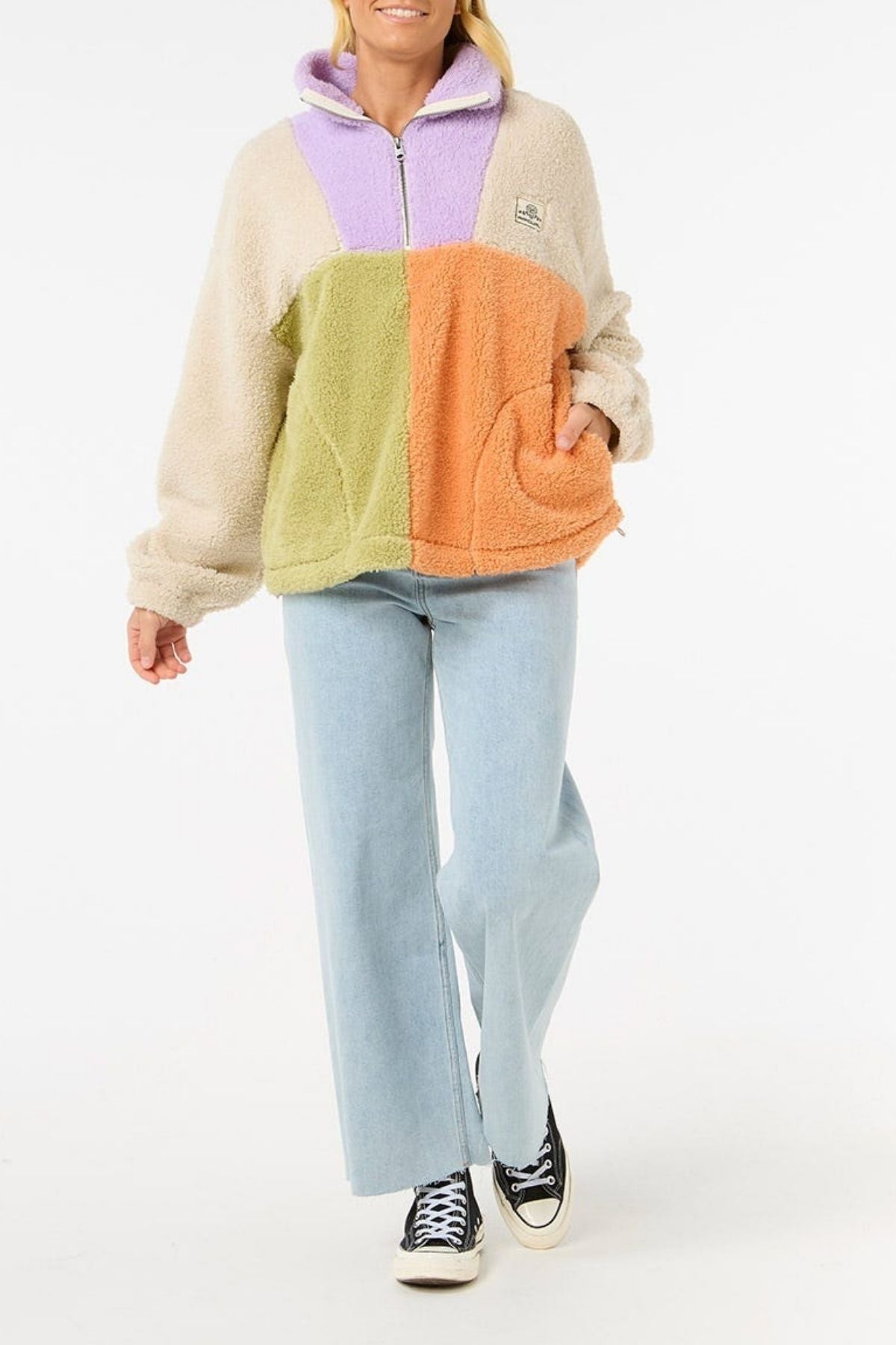 A person in the Rip Curl Block Party Polar Fleece with beige, purple, green, and orange color-blocked panels, paired with light blue jeans and black Converse sneakers, stands against a white background.