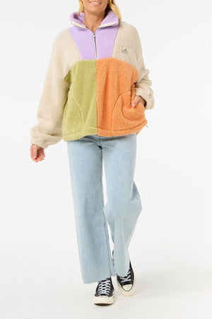 A person in the Rip Curl Block Party Polar Fleece with beige, purple, green, and orange color-blocked panels, paired with light blue jeans and black Converse sneakers, stands against a white background.