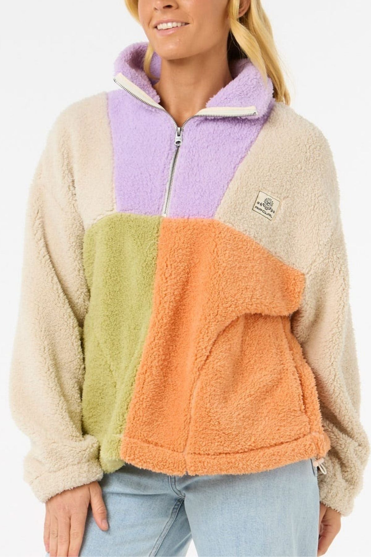 A woman wears the Rip Curl Block Party Polar Fleece, featuring beige sleeves with lavender, green, and orange panels. She pairs it with light blue jeans and smiles slightly.