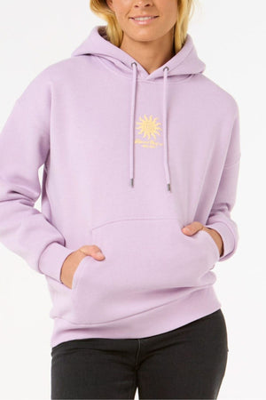 Wearing the Rip Curl Cala Heritage Hood—a light purple organic cotton-polyester fleece hoodie with a yellow sun graphic and chest text—a person stands hands-in-pocket against a plain white background.