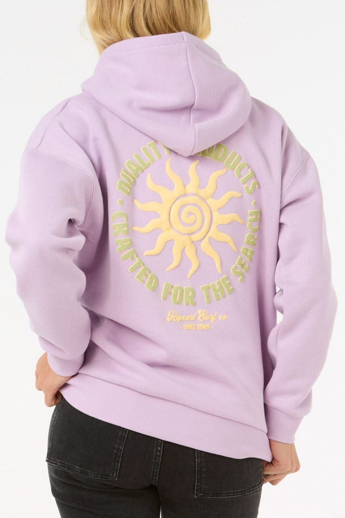 Wearing the Rip Curl Cala Heritage Hood in light purple, made from organic cotton-polyester fleece, with a yellow sun graphic and "Quality Products Crafted For The Search" on the back, in a heritage fit.