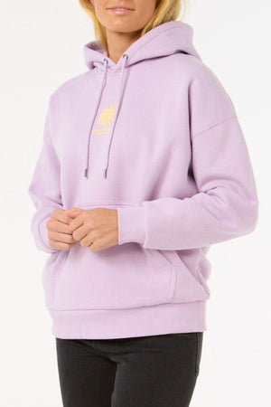 Someone wearing the Rip Curl Cala Heritage Hood in light purple, made from organic cotton-polyester fleece with a heritage fit and yellow sun chest graphic, pairs it with black pants against a plain white background.