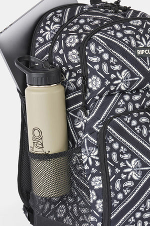 A black and white paisley print Rip Curl Chaser 33L Backpack with a computer tucked into the back compartment and a beige water bottle in the mesh side pocket.
