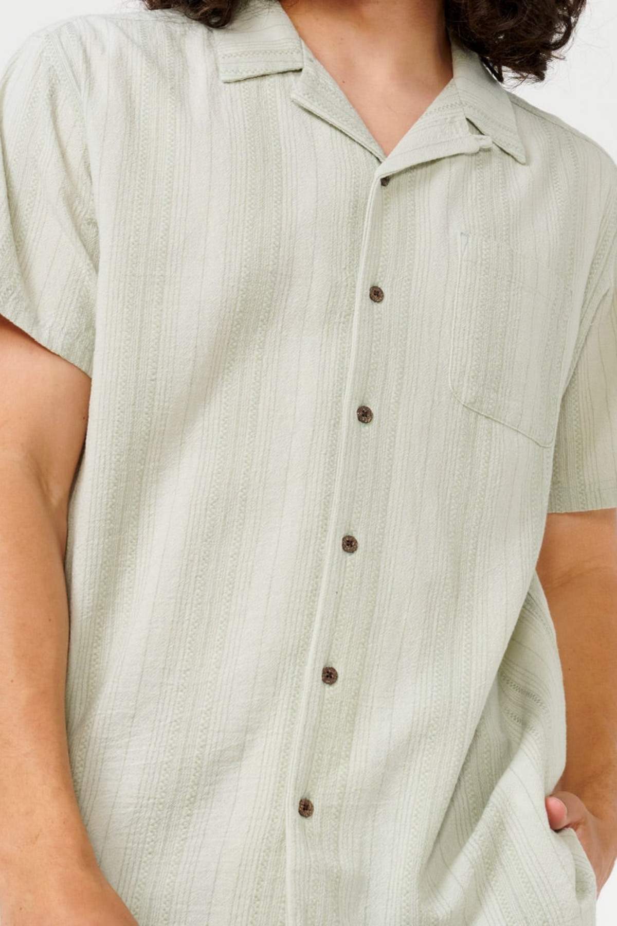 Someone is wearing the Rip Curl Check Mate Short Sleeve Shirt, featuring a light green color with vertical stripes. Made from cotton gauze for a relaxed Resort Fit, it has short sleeves, a button-up design, and a chest pocket. The person's left hand is in their pocket.