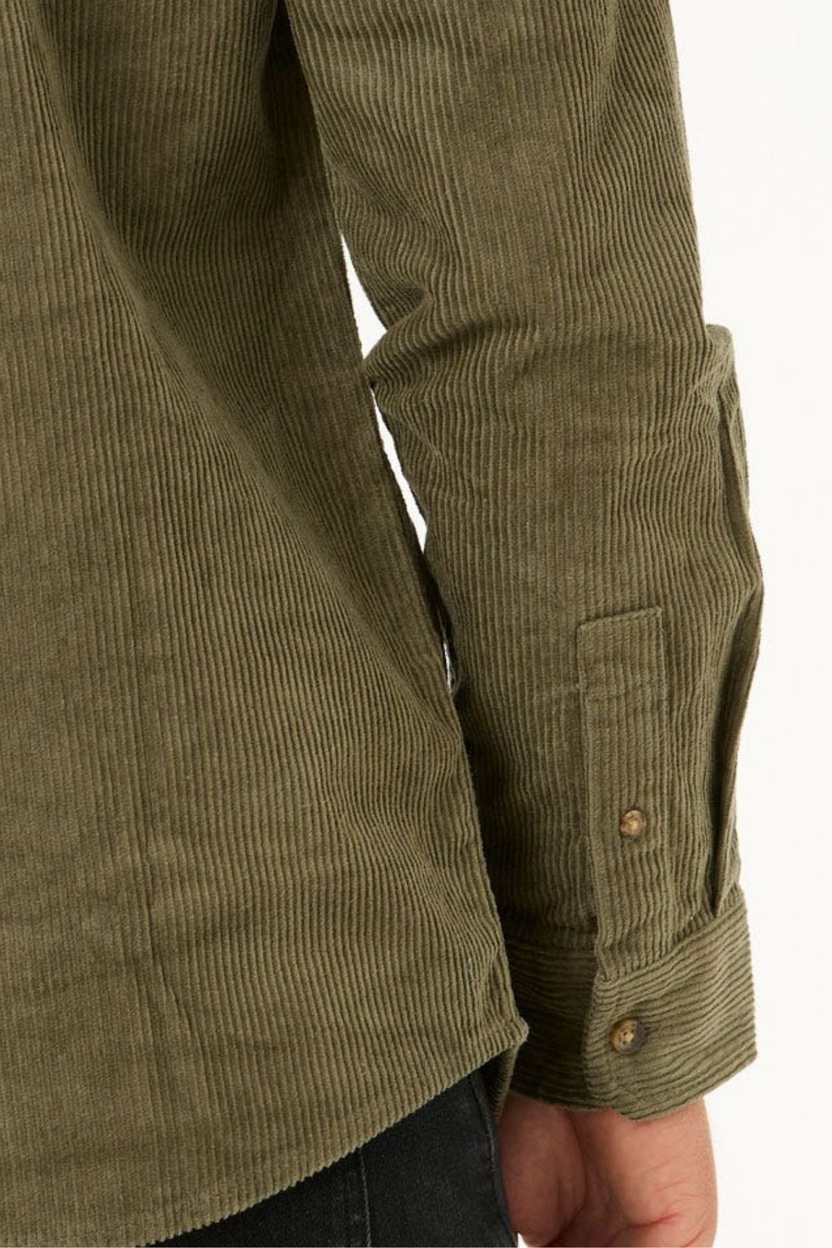 A close-up of a person’s arm wearing the Rip Curl Classic Surf Cord Shirt, an olive green long-sleeve cotton corduroy shirt with buttoned cuffs, next to black pants against a plain background.
