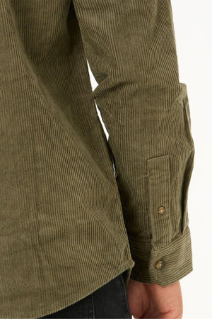 A close-up of a person’s arm wearing the Rip Curl Classic Surf Cord Shirt, an olive green long-sleeve cotton corduroy shirt with buttoned cuffs, next to black pants against a plain background.