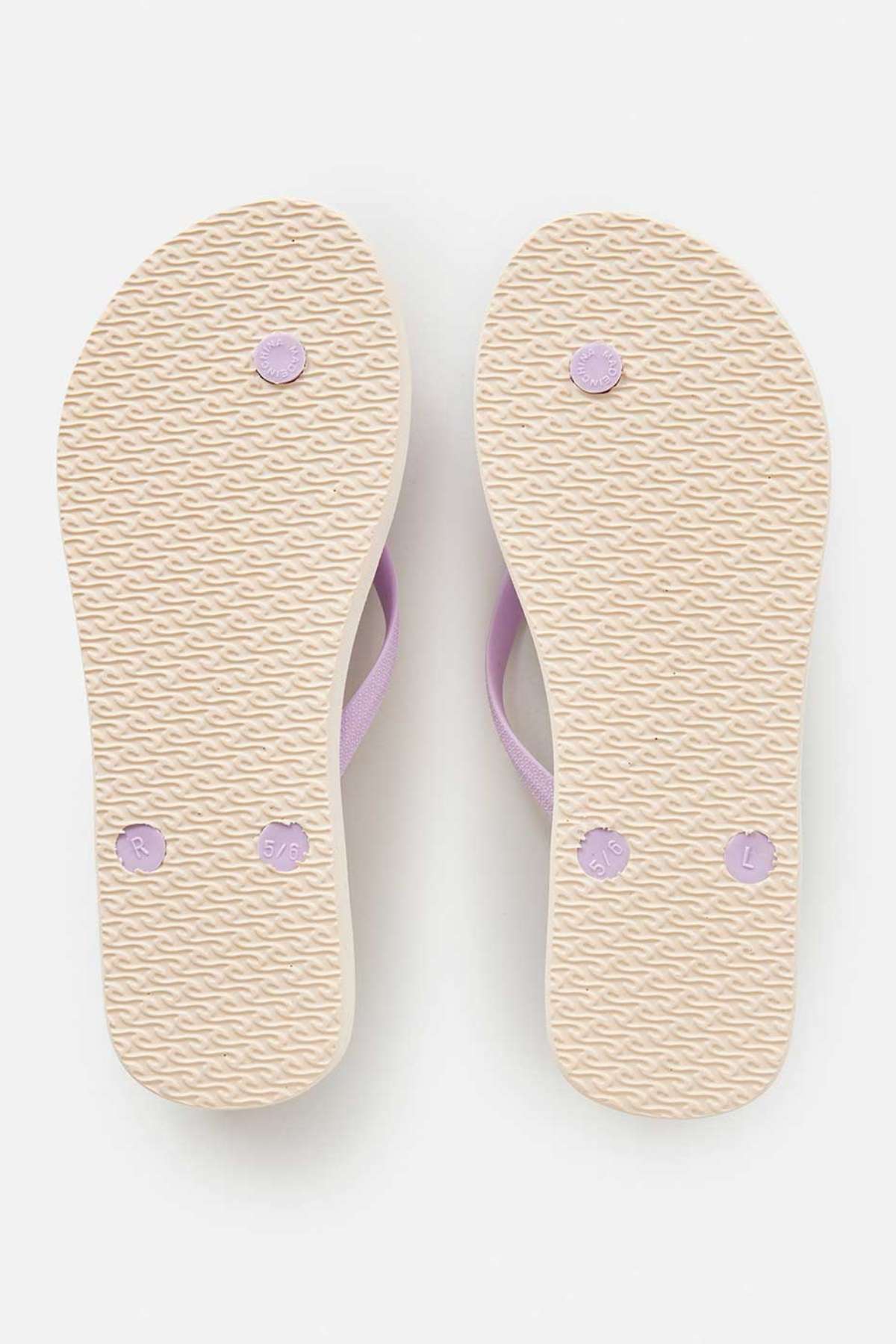 The image displays the bottom view of a pair of Rip Curl Classic Surf Bloom Thongs in beige, featuring BLOOM algae-blended foam for an eco-conscious design. The textured soles are complemented by subtle lavender accents, with one thong marked as size "5/6" and the other labeled "L." These sustainable flip-flops are elegantly positioned on a white background.