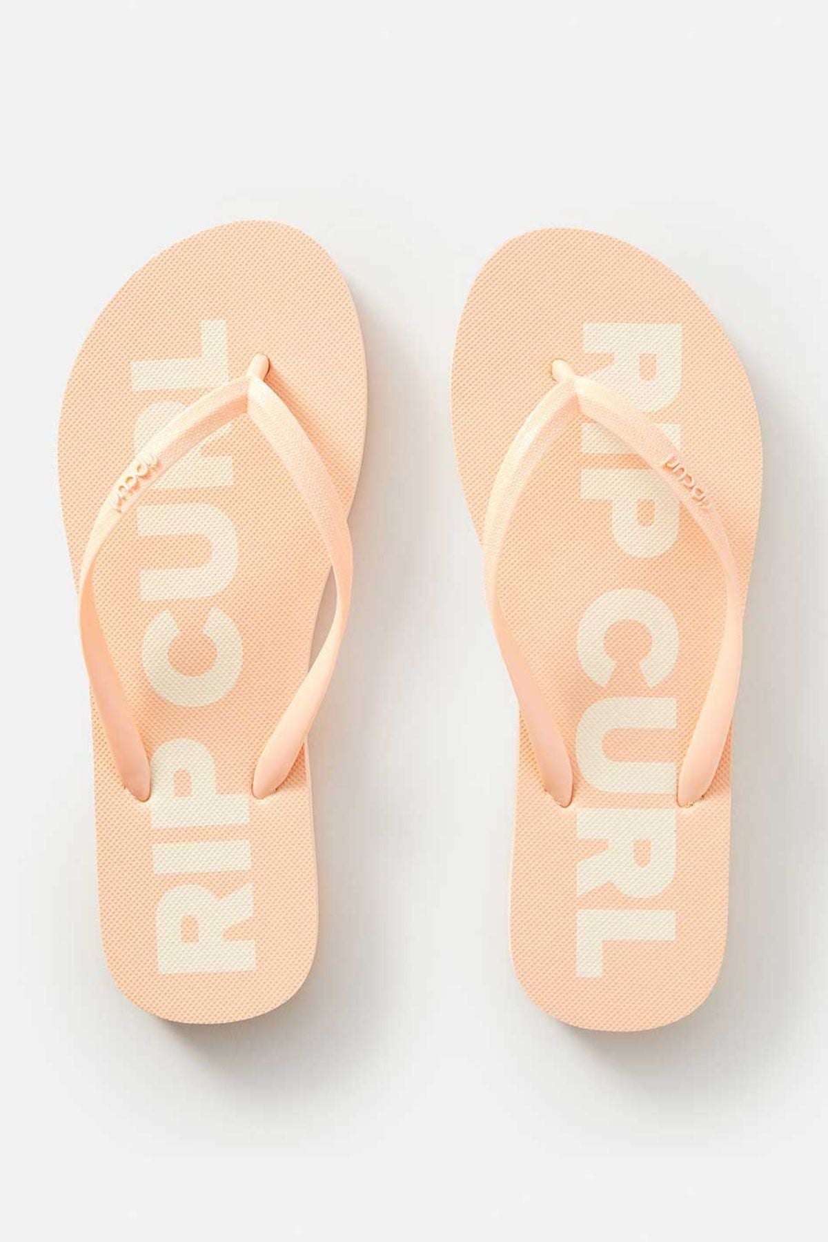 A pair of Rip Curl Classic Surf Bloom Thongs in peach, showcasing the prominent white "RIP CURL" text on the soles, made with BLOOM™ algae-blended foam EVA for eco-friendly comfort, presented against a light background.