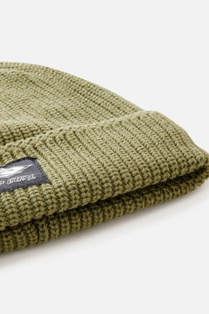 Close-up of the Rip Curl Classic Surf Lined Reg Beanie in olive green, made from soft cotton and folded at the edge. A black patch with a white logo and text decorates the front—perfect for your next outdoor adventure.
