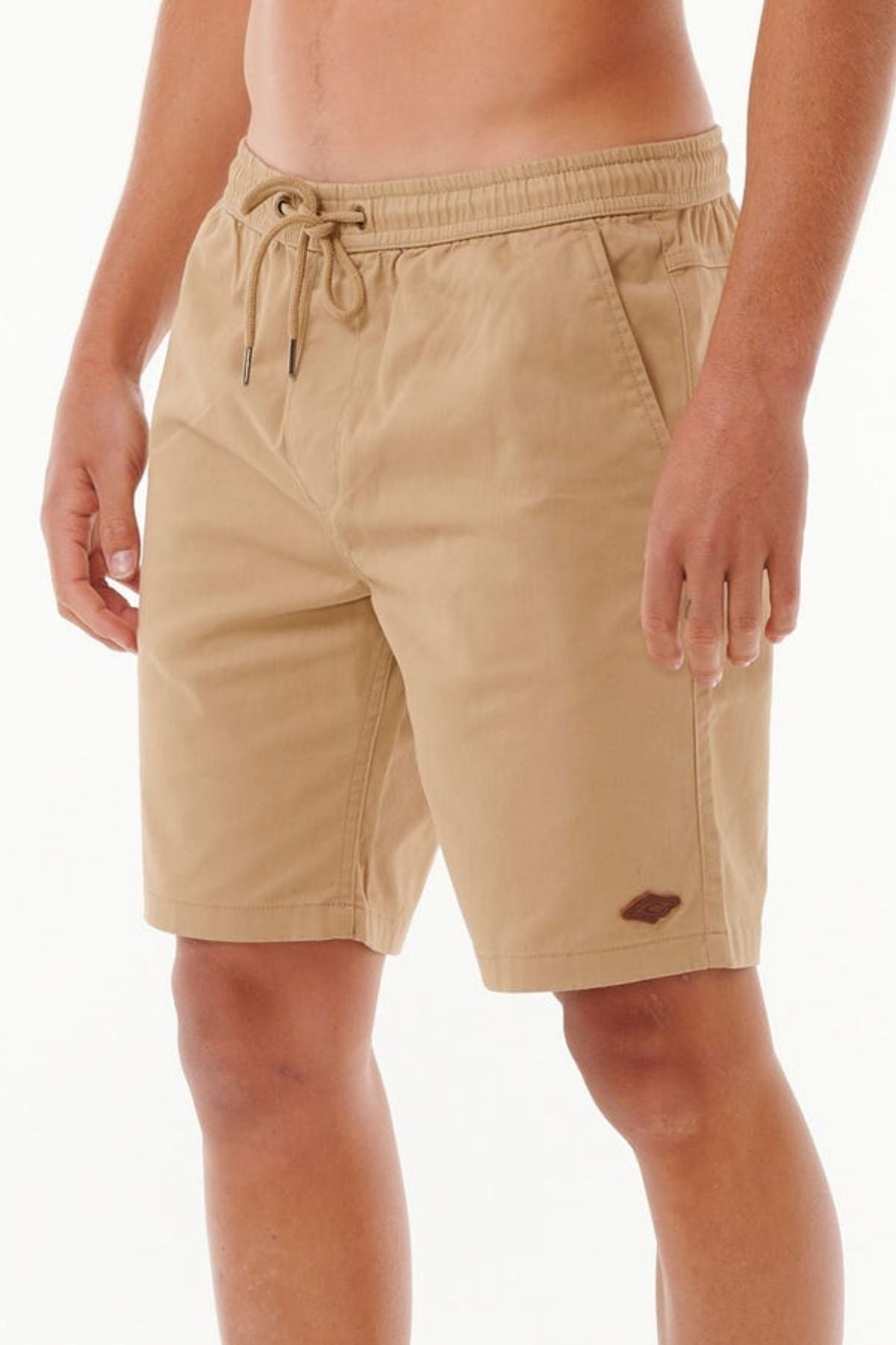 A person in Rip Curl Classic Surf Volley shorts, beige cotton with an elastic waistband and adjustable drawcord, poses against a white backdrop. The shorts have side pockets and a small brown logo on the left leg, with only their legs visible.