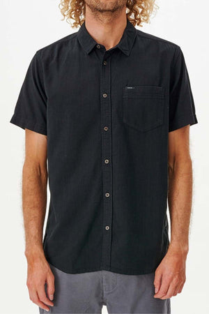 A person wearing the Rip Curl Classic Surf Washed Short Sleeve Shirt, a black cotton gauze button-up with a collar and chest pocket, stands with their arms relaxed by their sides. The shirt is paired with grey pants against a plain white background.