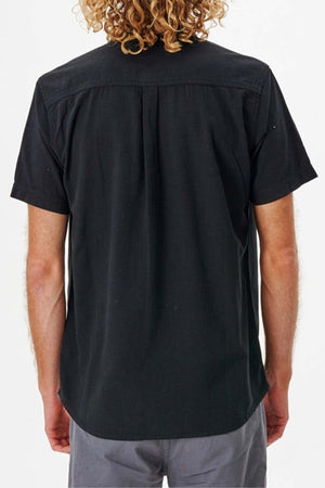 A person with curly hair is wearing a Rip Curl Classic Surf Washed Short Sleeve Shirt in black, paired with gray pants, viewed from the back against a plain white background.