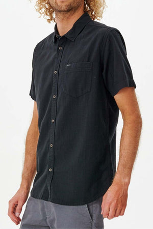 A person with curly hair dons a Rip Curl Classic Surf Washed Short Sleeve Shirt in black, crafted from cotton gauze, while standing against a plain background. The shirt includes a chest pocket and complements their gray pants exceptionally well.