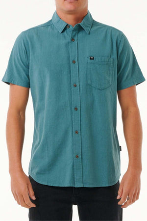 A person wearing the Rip Curl Classic Surf Washed Short Sleeve Shirt in teal, complete with a collar and a front pocket, is paired with black pants against a plain background. The shirt is distinguished by its brown buttons, enzyme wash finish, and a small tag on the pocket.