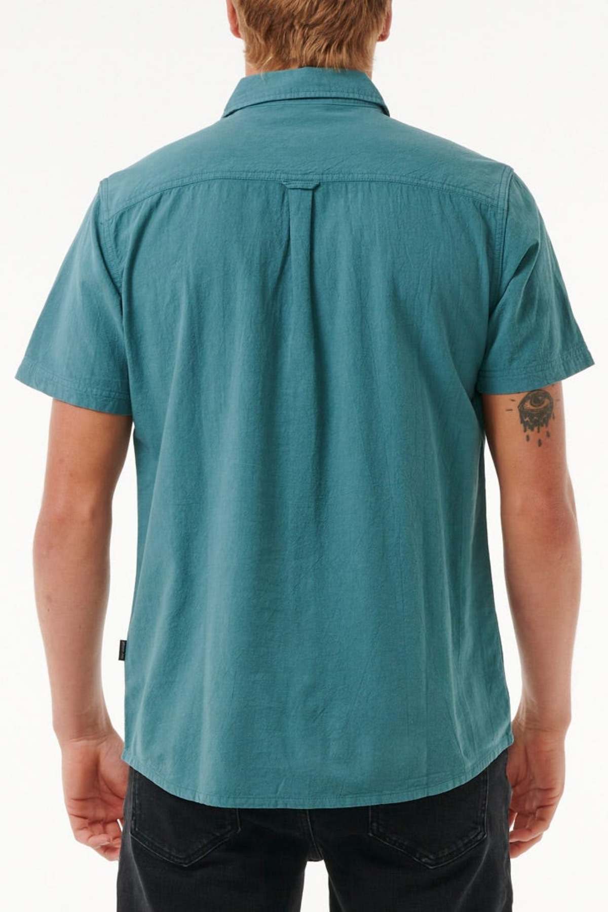 A person with short hair is wearing a Rip Curl Classic Surf Washed Short Sleeve Shirt in teal and black pants, facing away from the camera. They have a small tattoo on their right arm against a plain white background.