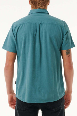 A person with short hair is wearing a Rip Curl Classic Surf Washed Short Sleeve Shirt in teal and black pants, facing away from the camera. They have a small tattoo on their right arm against a plain white background.