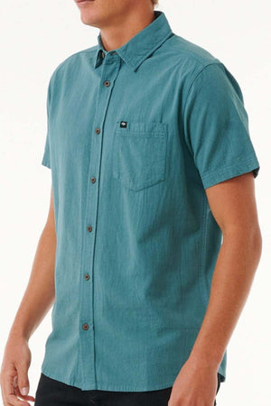 A person is wearing the Rip Curl Classic Surf Washed Short Sleeve Shirt in teal, made from soft cotton gauze. It features an enzyme wash for a relaxed, lived-in appearance and includes a chest pocket with a small logo patch. The background is plain and light-colored.