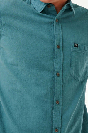 A close-up shows someone wearing the Rip Curl Classic Surf Washed Short Sleeve Shirt in teal, featuring a chest pocket with a small logo tag. The shirt is made from cotton gauze fabric with an enzyme wash finish and is accented with brown buttons. The person's face is not visible.