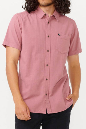 A person with long curly hair dons the Rip Curl Classic Surf Washed Short Sleeve Shirt—a short-sleeved, button-up pink shirt crafted from cotton gauze, featuring a chest pocket adorned with a small Rip Curl Classic Surf logo. They pair it stylishly with black pants, standing out against the plain white background.