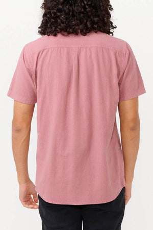 A person with curly hair is standing with their back to the camera, wearing a Rip Curl Classic Surf Washed Short Sleeve Shirt in mauve, which is crafted from soft cotton gauze paired with black pants. The plain white background accentuates the shirt's subtle pigment dye effect.