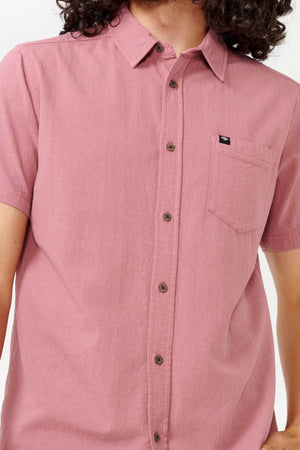 A person with long curly hair is wearing the Rip Curl Classic Surf Washed Short Sleeve Shirt, which is a button-up made from Cotton Gauze in a pink color. It features a pocket on the left side with a small black label. The image shows only their upper torso, without revealing their face.