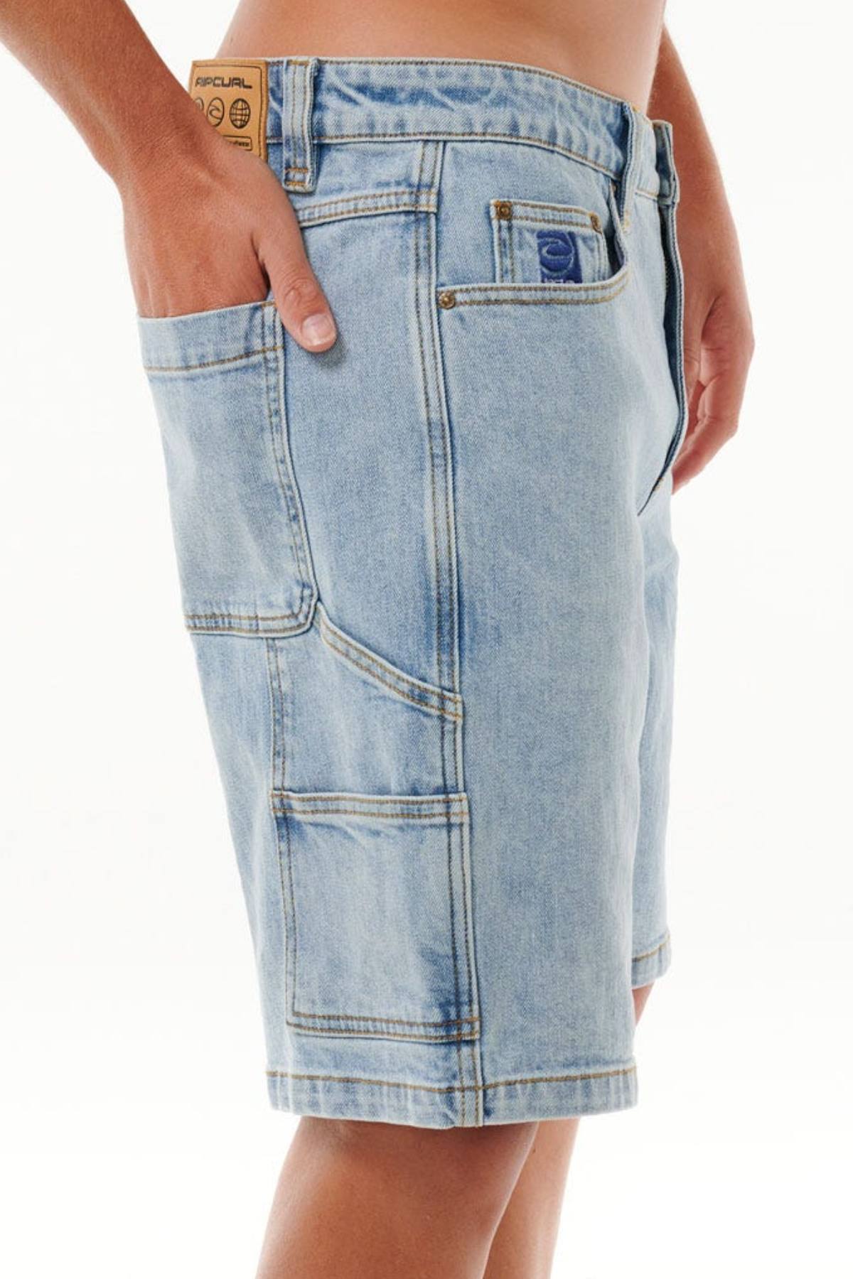A person wearing light blue Rip Curl Dosed Denim Walkshorts with multiple pockets, including a side pocket and a small label above the right front pocket, stands against a plain white background.